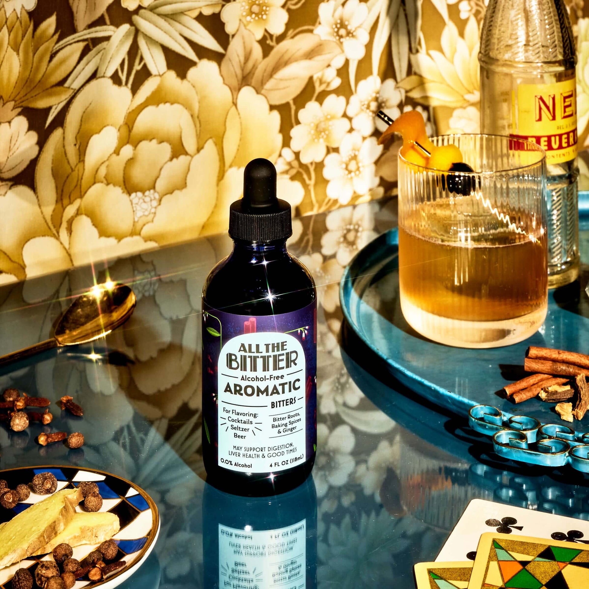 All The Bitter Aromatic non-alcoholic bitters pictured with a floral wallpapered background on a blue glass surface, a gold spoon, a cocktail in a whiskey glass on a turquoise tray with cinnamon sticks and other spices. Playing cards are pictured.